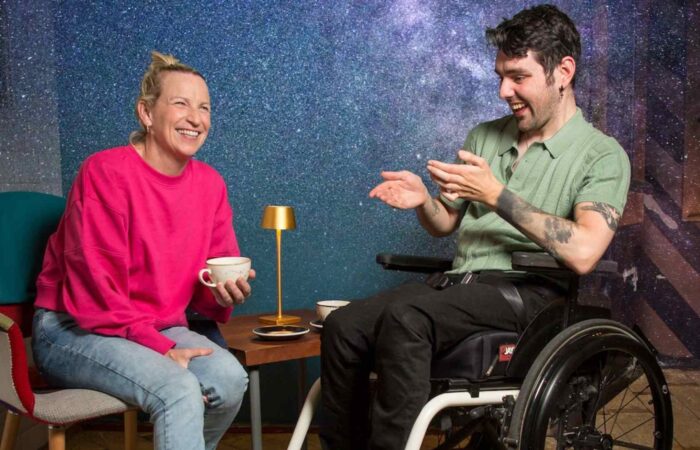 A photo of cast members Julie Sharkey & Eric Fitzgerald laughing. Julie is seated wearing a vibrant pink jumper and holding a cup of coffee. Eric, a wheelchair user, is dressed in a green polo shirt and appears to be telling Julie's character a funny story. The title No Magic Pill appears above their heads on a starry background