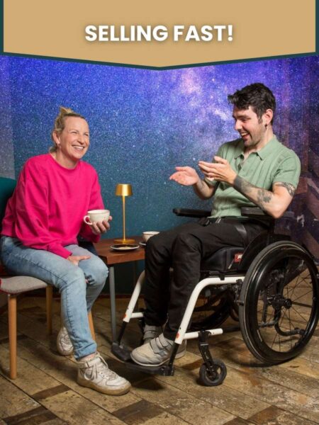 A photo of cast members Julie Sharkey & Eric Fitzgerald laughing. Julie is seated wearing a vibrant pink jumper and holding a cup of coffee. Eric, a wheelchair user, is dressed in a green polo shirt and appears to be telling Julie's character a funny story. The title No Magic Pill appears above their heads on a starry background