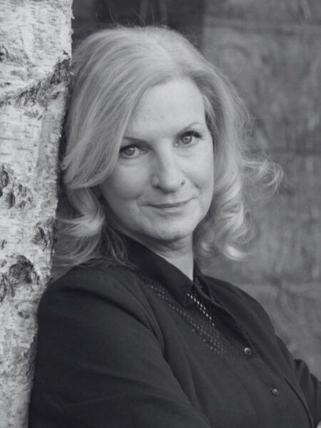 A monochromatic image of Eleanor Shanley leaning against a tree smiling at the viewer.