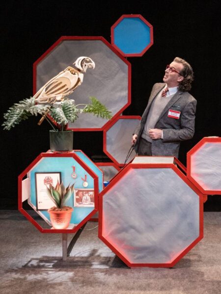 An official-looking man stands on a stage surrounded by octagonal props while talking to a mechanical parrot.