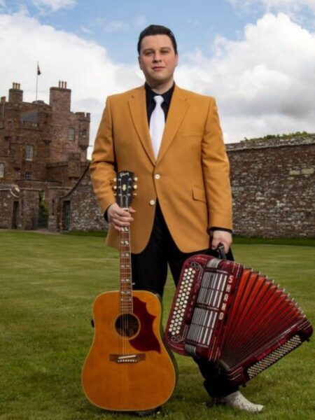 Daytime shot of singer Brandon McPhee standing on a grassy area in front of a Scottish castle holding an accordion and a guitar.