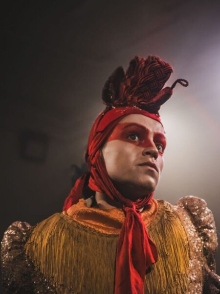 A woman dressed as a chicken with a band of red makeup across her eyes and a contemplative look on her face.