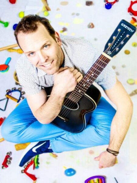 Kyle Riley sitting on a multicoloured floor holding a guitar, smiling while glancing up at the viewer.