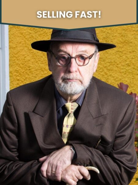 Michael Harding, wearing a trilby hat, slightly stooped while holding a cane, standing against the yellow-painted wall of a house. The words "selling fast" are overlaid above.