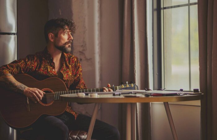 Niall McCabe sitting in a dimly-lit room playing guitar in front of a window