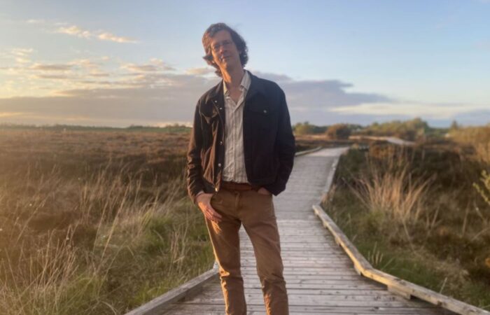 Manchan Magan standing on a wooden path in a bog-land location as promotion for Rewilding the Mind