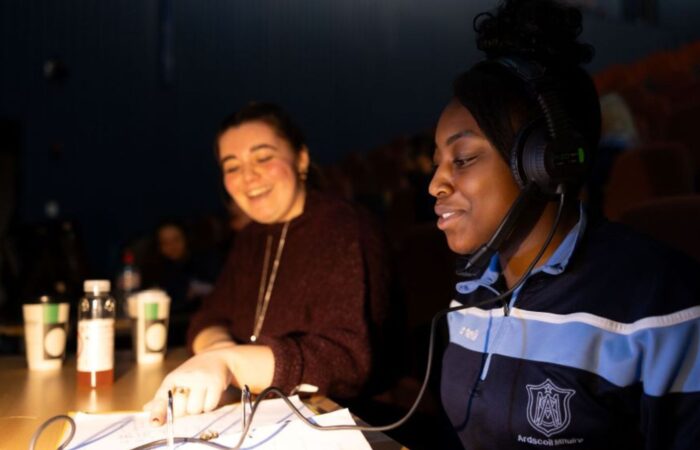 Two young women, one wearing a headset and microphone, sitting at a sound desk reading a script for Women in Technical Theatre