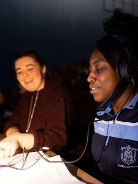 Two young women, one wearing a headset and microphone, sitting at a sound desk reading a script for Women in Technical Theatre