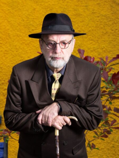 Michael Harding, wearing a trilby hat, slightly stooped while holding a cane, standing against the yellow-painted wall of a house