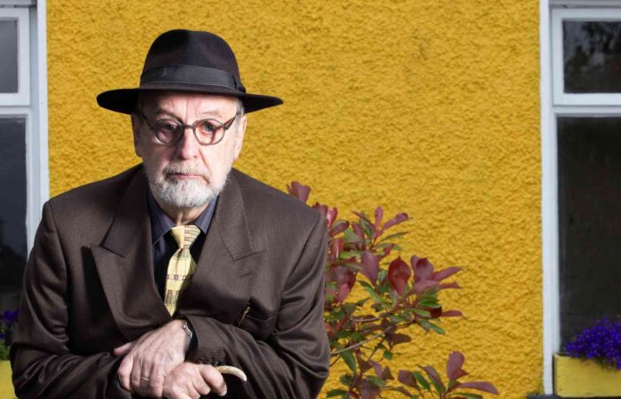 Michael Harding, wearing a trilby hat, slightly stooped while holding a cane, standing against the yellow-painted wall of a house