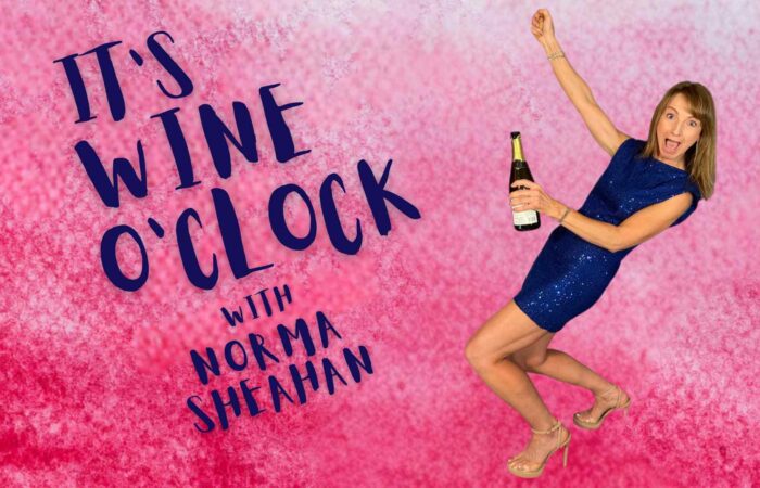 Norma Sheahan wearing a blue cocktail dress, leaning back while popping the cork on a bottle of champagne with her right hand raised in the air and a mischievous look on her face, set against a dappled pink background overlaid with the words "It's Wine O'Clock with Norma Sheahan."