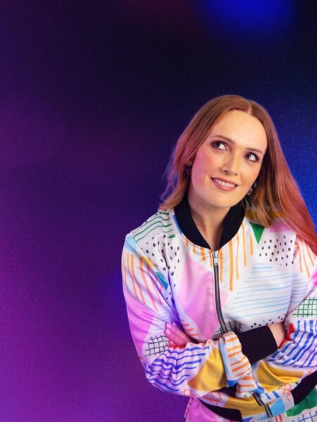 Comedian Emma Doran wearing a multicoloured jacket, posing with her arms crossed, her head slightly tilted, set against a purple-blue background. She is smiling as she looks off to her right with a whimsical look on her face.