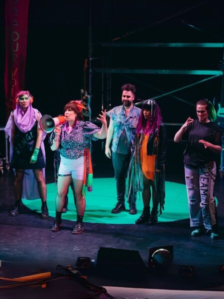 A diverse group of performers from Punk Alley - wearing punk-style garb - are standing on a stage. One of the female performers is shouting into a megaphone.