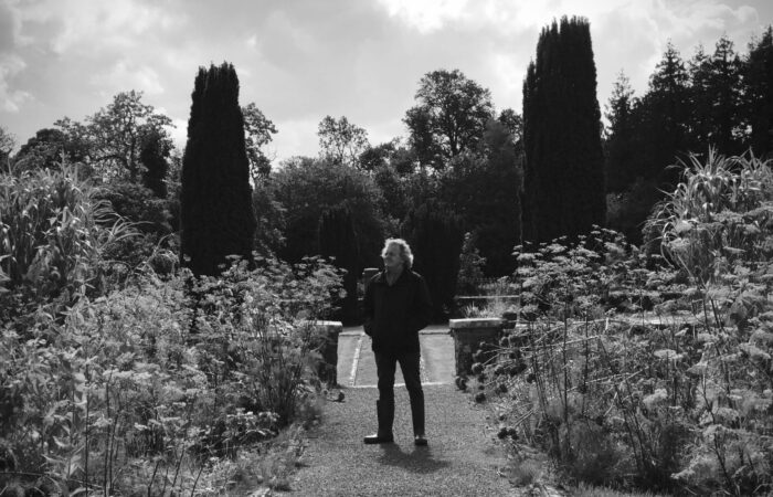 Monochromatic image of an older man with his hands in his pockets standing in a garden surrounded shrubbery.