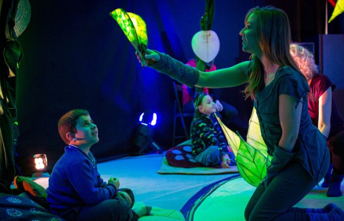A young, smiling boy wearing a blue jumper sits on the floor smiling up at a woman holding a glowing leaf over his head. Other children can be seen in the background. The show is Lullaby Leaf.