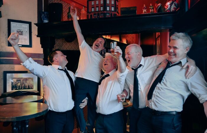 The Kings of the Kilburn High Road: Five men in white shirts and ties celebrate joyfully in a pub, smiling and holding drinks. The atmosphere is lively and cheerful.