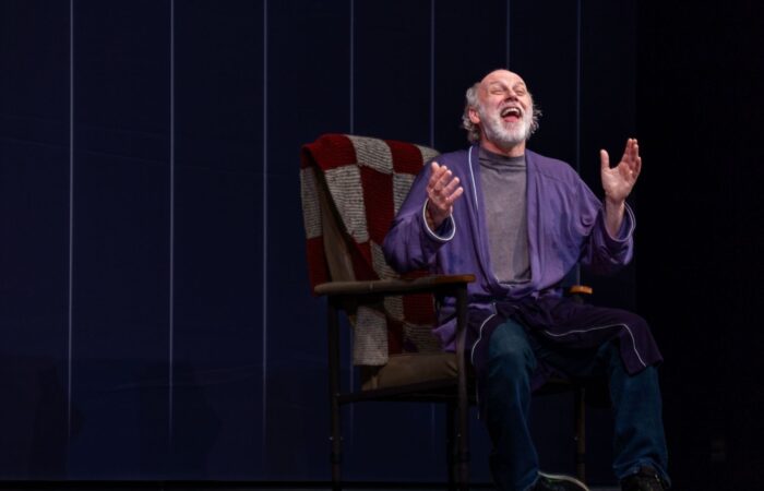 A man wearing pajamas sits in a chair laughing heartily, raising his hands in a gesture of joy and excitement in The Life and Times of Paddy Armstrong.