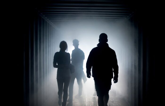 Three people in silhouette in a darkened tunnel with a bright white light behind them. All Things.