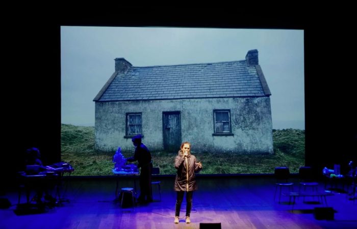 Aliens - A woman stands at a microphone on a dark theatre stage, with a large projection of an isolated weathered cottage in a grassy landscape behind them. Other performers and tables of props are dimly lit at either side of the stage under blue lighting.
