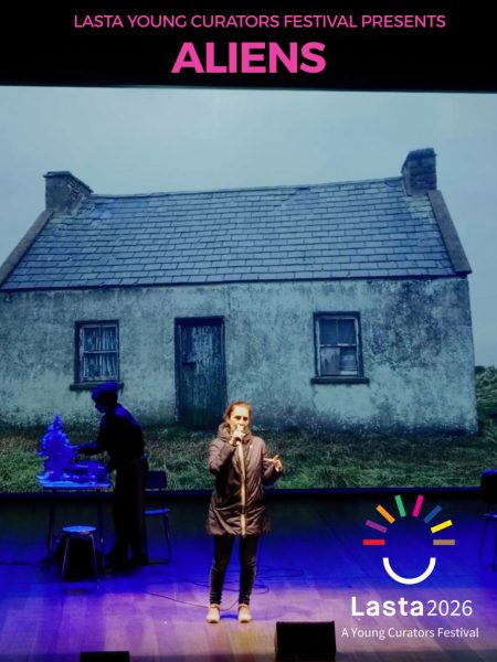Aliens - a woman stands centre stage speaking into a microphone while another figure works at a table in shadow to the left. Pink text at the top reads "LASTA Young Curators Festival Presents: ALIENS", with the Lasta 2026 festival logo displayed in the bottom-right corner.
