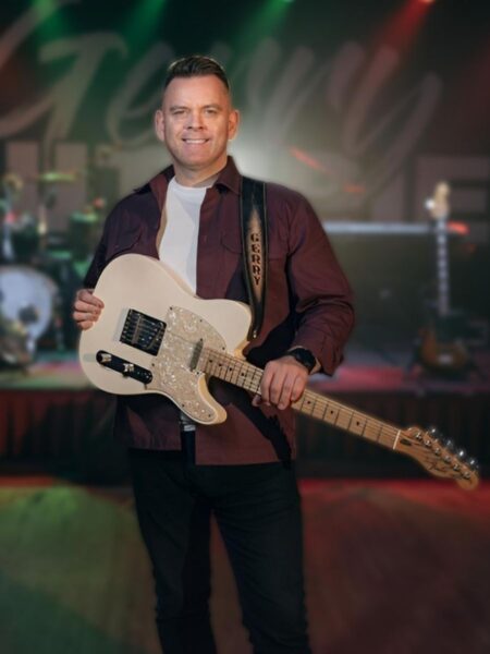 Gerry Guthrie holds an electric guitar on stage, with colorful stage lights and blurred band equipment in the background, conveying a lively concert vibe.