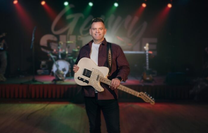 Gerry Guthrie holds an electric guitar on stage, with colorful stage lights and blurred band equipment in the background, conveying a lively concert vibe.