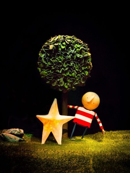How To Catch A Star - A puppet in a red-and-white striped shirt reaches towards a large yellow star beside a leafy topiary tree on a grass-covered stage, under dramatic theatrical lighting.