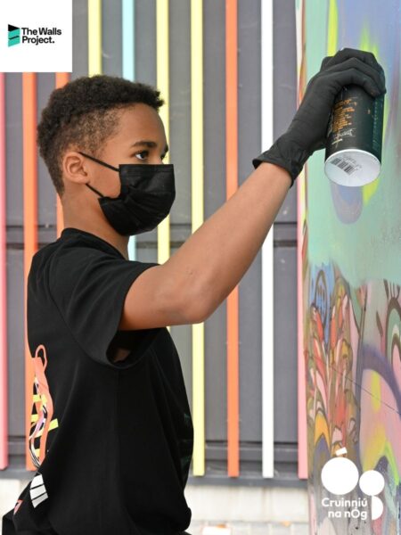 Wonder Walls - A young artist wearing a black mask and gloves spray paints a colorful mural. The scene is vibrant, with lively hues of pink, blue, and yellow.