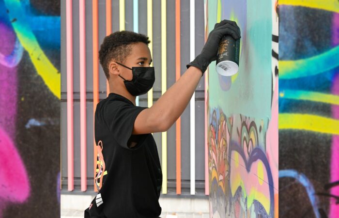 Wonder Walls - A young artist wearing a black mask and gloves spray paints a colorful mural. The scene is vibrant, with lively hues of pink, blue, and yellow.