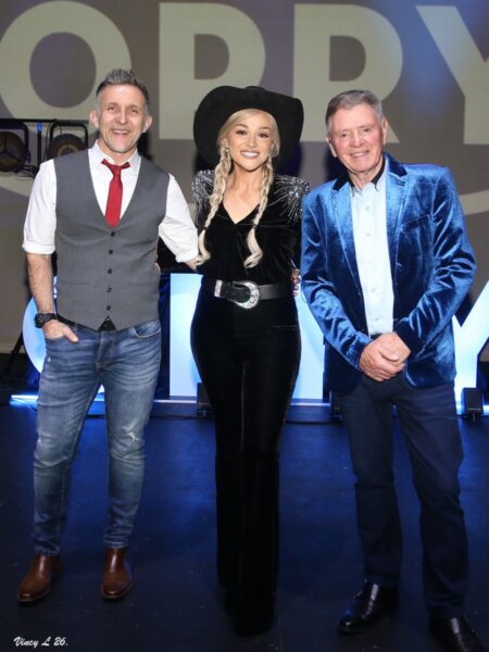 A Night at the Opry - Three performers pose together on stage, smiling at the camera. The person in the center wears a black cowboy hat and black outfit with silver detailing and braided hair, while the people on either side wear smart stage attire, including a grey waistcoat with a red tie and a bright blue velvet jacket. Stage lighting and a large projected backdrop create a concert-style atmosphere.