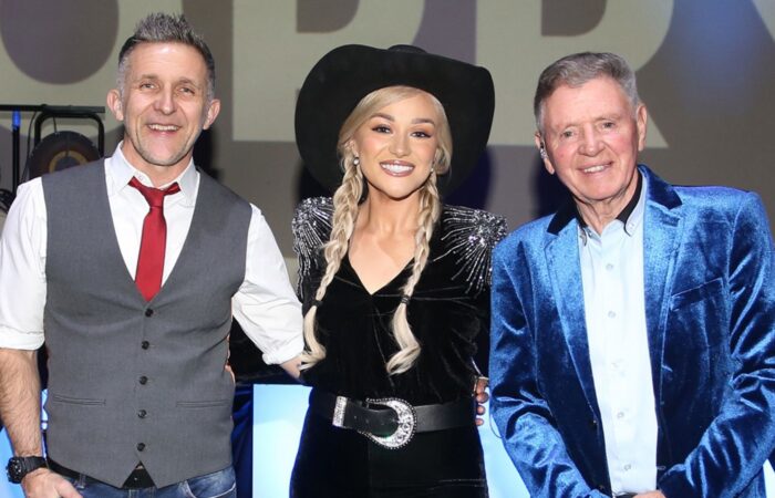 A Night at the Opry - Three performers pose together on stage, smiling at the camera. The person in the center wears a black cowboy hat and black outfit with silver detailing and braided hair, while the people on either side wear smart stage attire, including a grey waistcoat with a red tie and a bright blue velvet jacket. Stage lighting and a large projected backdrop create a concert-style atmosphere.