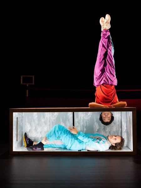 Flip - Two contemporary dancers perform a playful balancing act on a dark stage. One dancer lies inside a long, box-like prop wearing a turquoise tracksuit, while the other balances upside down on top of the box in bright purple trousers and a red shirt, creating a striking, whimsical image that suggests movement, creativity and connection.