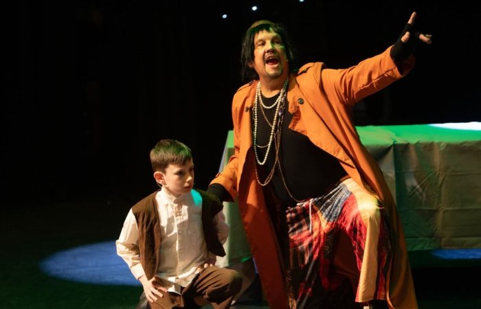 Longford Variety Group: A child in a white shirt and brown vest stands beside an adult performer on a dimly lit stage. The adult, wearing an orange coat, layered necklaces, and a colorful skirt, kneels and points dramatically into the distance while speaking or singing. Stage lighting highlights the performers against a dark background, with a covered table visible behind them.