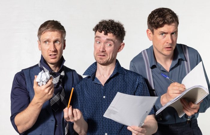The three members of Foil Arms and Hog stand against a light grey background with exaggerated, puzzled expressions while holding notepads, paper, and pencils, suggesting a comedy writing session.