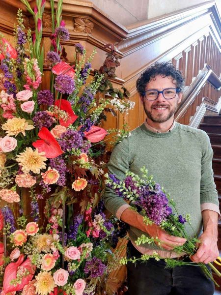 Longford Flower Club Christmas Gala 2026 - A smiling florist stands beside a large, colourful floral installation in an elegant wood-paneled interior with a grand staircase. He holds a bouquet of fresh purple flowers, while the display features pink roses, red anthuriums, purple alliums, gladioli and other seasonal blooms arranged in a tall, dramatic design.