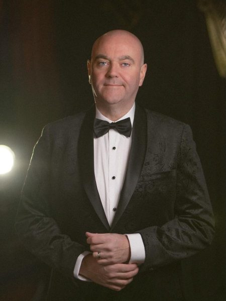 Patrick Feeney stands in a black tuxedo with a bow tie with his hands clasped, illuminated by stage lighting against a dark background.