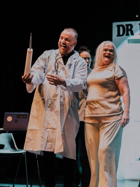 Longford Variety Group: Two performers stand on stage during a comedic theatre scene. One wears a white lab coat and holds up a large syringe prop while laughing, as another performer in light-coloured clothing smiles beside them. A laptop, stage set, and clinic-style backdrop are visible in the background, adding to the humorous medical-themed performance.